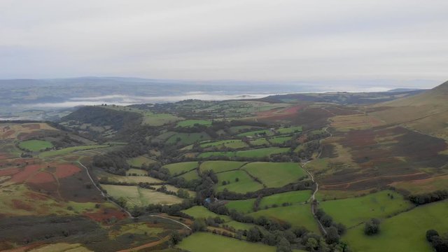 An aerial backward footage of a Welch valley at dusk with green pasture, trees, rolling hills under a cloudy and misty sky 