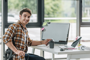Naklejka na ściany i meble happy programmer smiling at camera while sitting near computer monitor in office