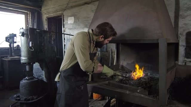 Pan shot of Caucasian bearded blacksmith in apron and gloves approaching to stove with metal billet in his hands, putting it in coals and moving stove damper to blow fire up