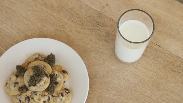 Pan across cookies and milk and cannabis buds on wooden table and white plate