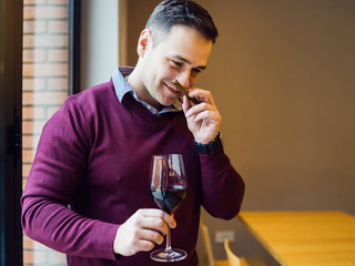 Young man wearing purple sweater holding a glass of red vine and smelling cigar at home office or party at restaurant hotel