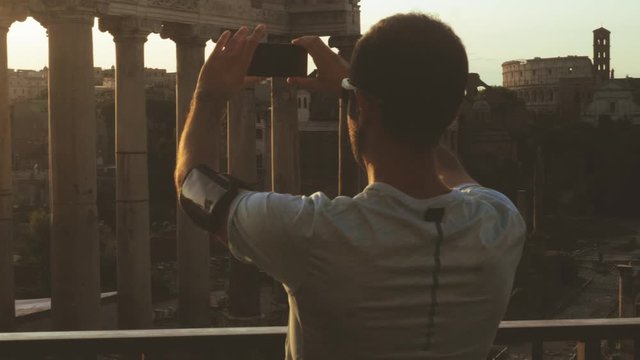 Young man in running clothes taking pictures in front of the Roman Forum at sunrise. Historical imperial Foro Romano in Rome, Italy from panoramic point of view.