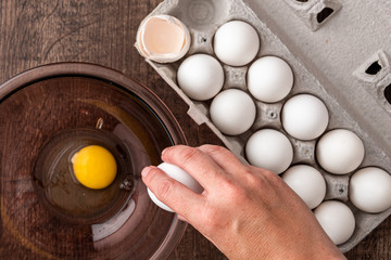Eggs in cardboard egg carton, woman’s hand breaking egg on side of glass bowl, raw egg in bowl,...