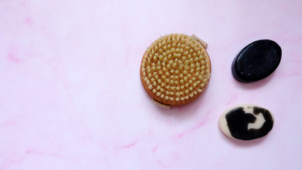 A round body scrub brush with two bars of black charcoal and milk body soap, on pink surface.