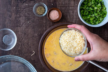 Ingredients for quinoa egg bake, raw egg mix in glass bowl with metal whisk, quinoa, cheese, herbs, broccoli, glass baking dish, on a wood table