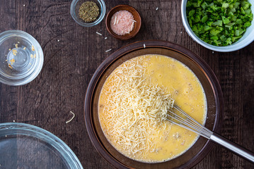 Ingredients for quinoa egg bake, raw egg mix in glass bowl with metal whisk, quinoa, cheese, herbs, broccoli, glass baking dish, on a wood table