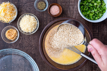 Ingredients for quinoa egg bake, raw egg mix in glass bowl with metal whisk, quinoa, cheese, herbs, broccoli, glass baking dish, on a wood table