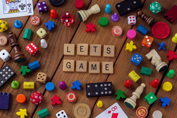 "Lets Game" spelled out in wooden letter tiles, surrounded by dice, cards, dominoes, chess pieces and other game pieces on a wooden background
