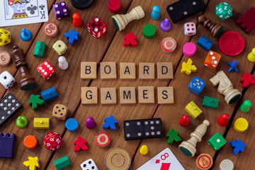 "Board Game" spelled out in wooden letter tiles, surrounded by dice, cards, dominoes, chess pieces and other game pieces on a wooden background