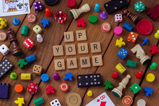 "Up Your Game" spelled out in wooden letter tiles, surrounded by dice, cards, dominoes, chess pieces and other game pieces on a wooden background