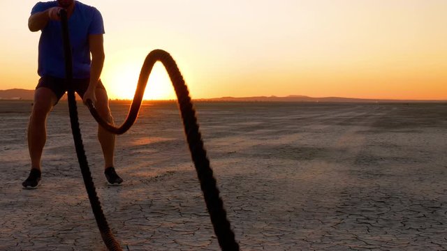 Athletic man working out with battle ropes on a dry lake at sunset