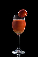 a glass of tomato juice stands on a reflective surface on a dark background and a round red tomato hangs on the neck.