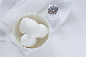 four white eggs lie in a plate on a white wooden table next to a towel and salt shaker.