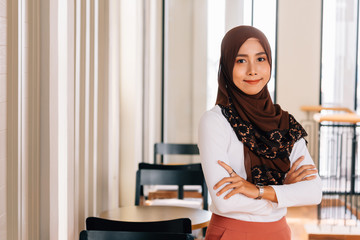 Young happy and successful South East Asian Islamic business woman with arms crossed in business corporate building setting looks at camera. She wears head scarf or hijab and modern elegant clothes