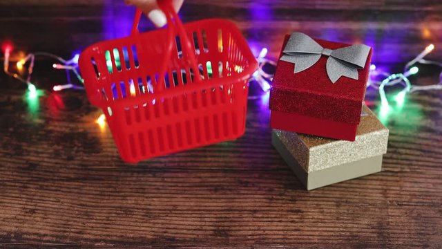 Xmas presents concept hand placing shopping basket next to festive Christmas gift boxes with fairy lights background