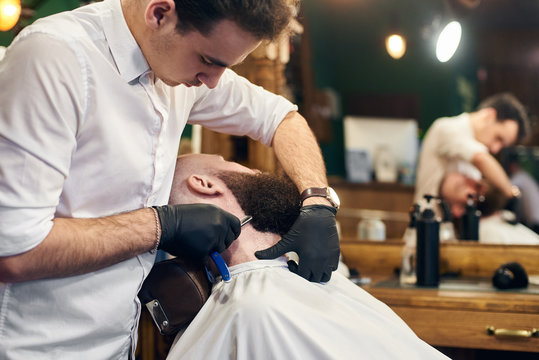 Man client reclining in chair for shaving by barber hands in gloves with sharp blade on bearded face. Trust, skillful hands, servicing, barbershop concept. Blurred background
