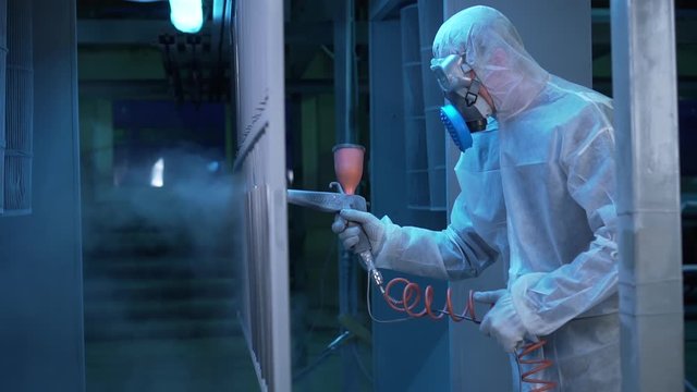 Industrial production, metal painting shop, special dark lighting in room, worker in special suit is spraying hazardous paint, small particles in air, pulverizer, breathing mask with cleaning filter
