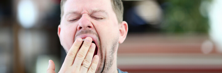 Obraz premium Young attractive man yawns in cafe covering mouth with hand. Syndrome of chronic lack of sleep due to processing overwork concept.