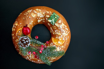 Christmas round fruit cake decorated with fir tree branch, glazed fruits and nuts over  black background. Roscon de reyes. King cake Top view. Copy space