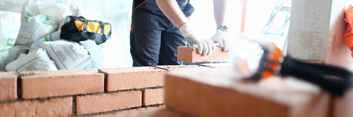Male builder hand in gloves holding clay brick on background of brickwork wall. Repair and...