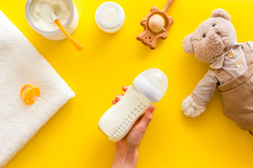 Milk bottle for feed baby in hands near toys and accessories on yellow background top view pattern