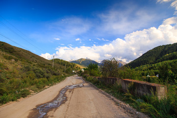 Country road high in the mountains. Tall trees, snowy mountains and white clouds on a blue sky. Kyrgyzstan Beautiful landscape.