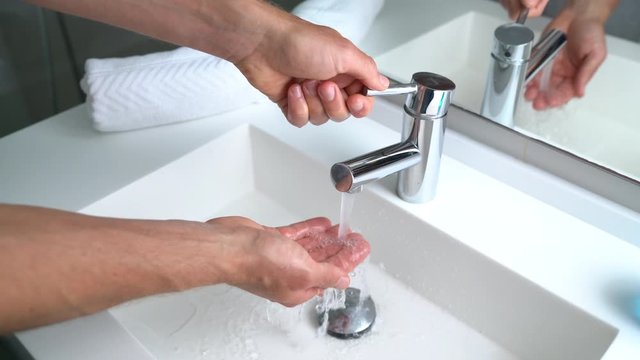 Man washing hands in bathroom sink at home checking temperature touching running water with hand. Closeup on fingers under hot water out of a faucet of a sink.