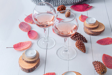 Small candles, two glasses with rose wine, cones, dry red leaves, gray scarf knitted on a white wooden table. Hello, Autumn. Cozy autumn background.