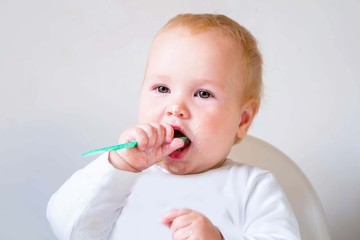 Baby, little girl eating lunch with green plastic spoon. Delicious food. Baby eating with pleasure. White plastic chair for feeding.