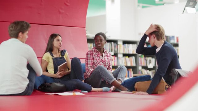 Group of four college students, two boys and two diverse girls, laughing while talking on large sofa in lounge area of library