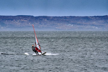 A male athlete is interested in windsurfing. He moves on a Sailboard on a large lake on an autumn day.