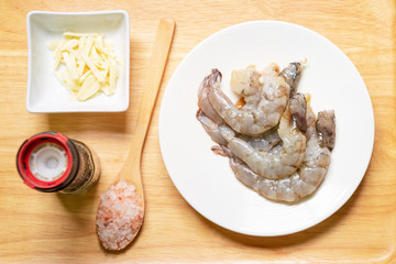 Peeled shrimps in a white plate with slice garlic, black pepper and Himalayan pink salt in wooden spoon on table prepare for cook. Top view