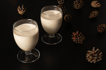 Eggnog Christmas cocktail in a transparent wine glass, ready to be sprinkled with cinnamon and nutmeg, on a black background with Pine cones.