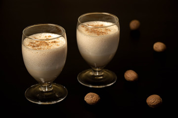 Eggnog Christmas cocktail in a transparent glass, sprinkled with grated nutmeg, on a black background with whole nutmeg.