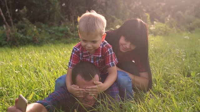 happy family teamwork outdoors have fun concept outdoors slow motion video. mom dad and son take a photo with a smartphone in nature are sitting on the grass have fun playing. mom girl dad man