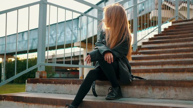 Teenager girl in black leather jacket sitting on stairs on urban landscape. Stylish girl in black leather jacket sitting on staircase looking away