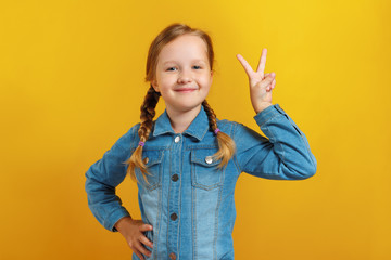Cheerful little girl shows a victory sign. A child in a denim shirt on a yellow background. Success...