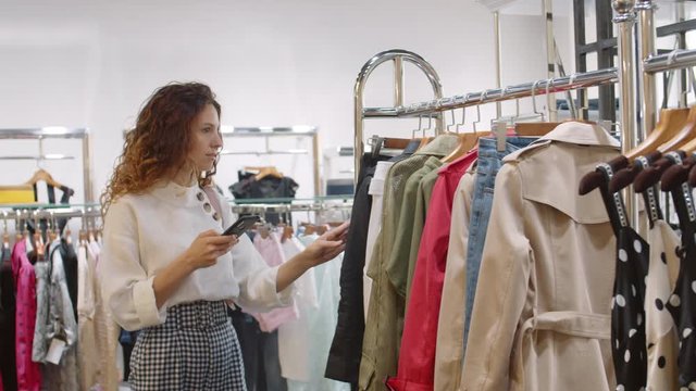Medium shot of Caucasian woman with long curly hair with smartphone in her hand choosing clothes in shop