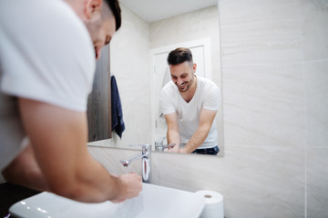 Handsome caucasian man in pajamas washing his face in washbasin in morning. Bathroom interior.