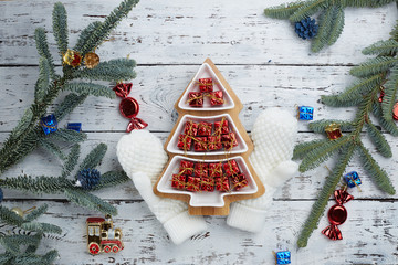 lots of little red gift boxes in the shape of a Christmas tree on a white wooden background. White knitted mittens. Fir branches on the edge of the frame . The concept of Christmas composition.