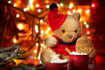 Christmas composition. A red mug with marshmallows stands in front of a Teddy bear in a red cap on the background of the lights of the Christmas garland. brown wooden background