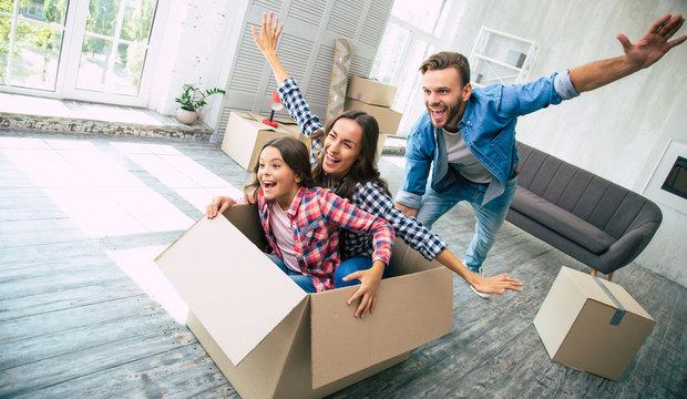 Family of three is playing in their new apartment after the house moving, young bearded father is riding his wife and daughter in a box, which was just emptied from their personal items.