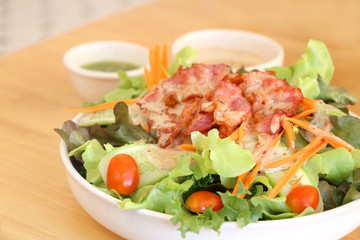 Close up fresh vegetable salad (lettuce, carrot, cucumber, tomato)  with fried bacon and roasted sesame Japanese dressing in white bowl on wooden table background