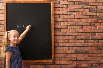 Cute little left-handed girl doing sums on chalkboard near brick wall