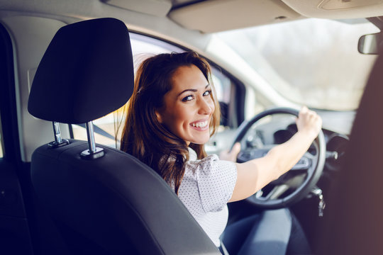 Rear view of smiling caucasian brunette dressed smart casual driving her expensive car while looking at camera. Hands are on steering wheel.