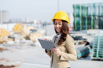 Attractive smiling Caucasian female architect in beige jacket and with protective helmet on head...