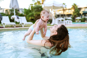 Fototapeta na wymiar Charming Caucasian brunette lifting her 6 months old son while standing in swimming pool. First time at pool concept.