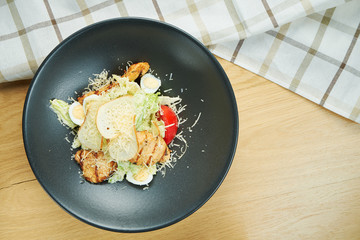 Caesar salad with croutons, parmesan, chicken, egg in a black stylish bowl. Restaurant serving. Selective focus, wooden background