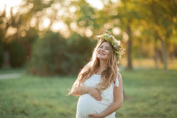 Portrait of a pregnant woman in the sun. Young beautiful pregnant woman with a wreath on her head in the field. Motherhood. Warm autumn.
