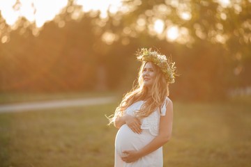Fototapeta na wymiar Portrait of a pregnant woman in the sun. Young beautiful pregnant woman with a wreath on her head in the field. Motherhood. Warm autumn.